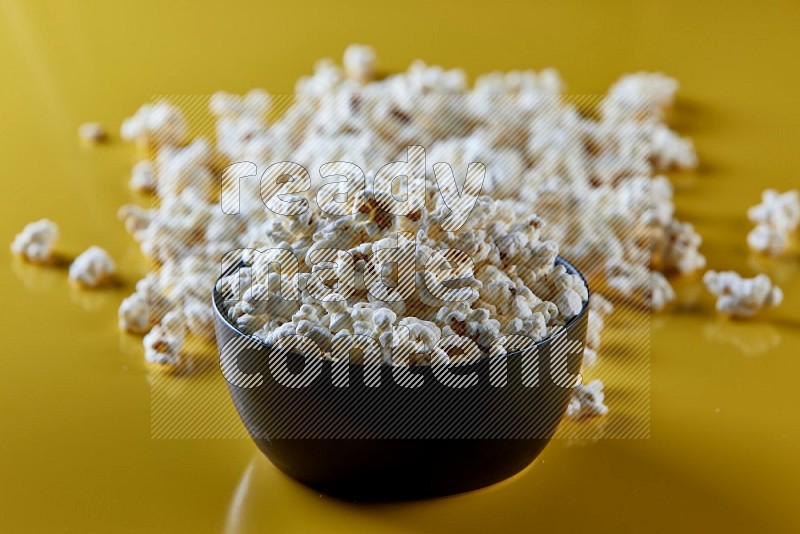 A black ceramic bowl full of popcorn with popcorn beside it on a yellow background in different angles