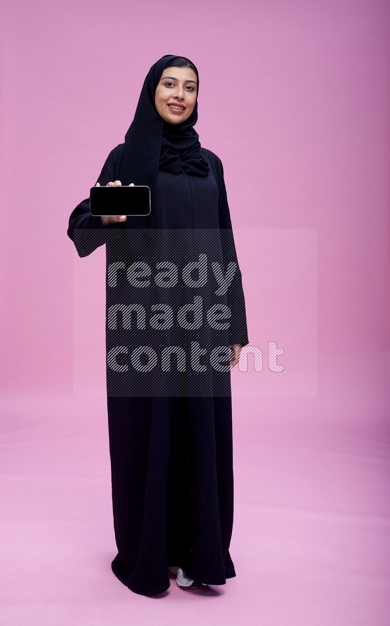 Saudi woman wearing Abaya standing showing phone to camera on pink background