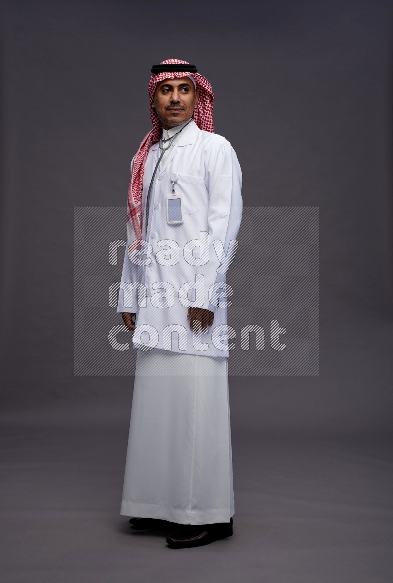 Saudi man wearing thob with lab coat and shomag with pocket employee badge with stethoscope standing interacting with the camera on gray background