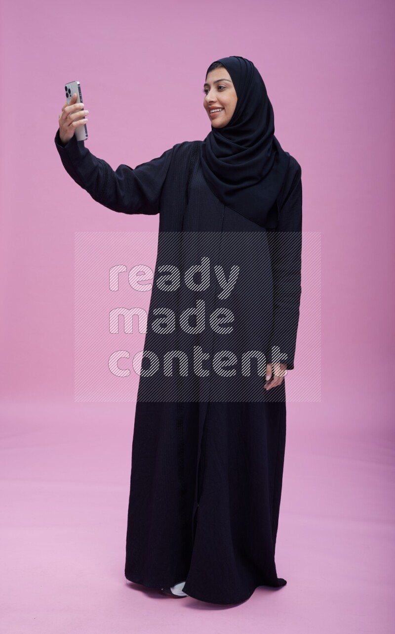 Saudi woman wearing Abaya standing taking selfie on pink background