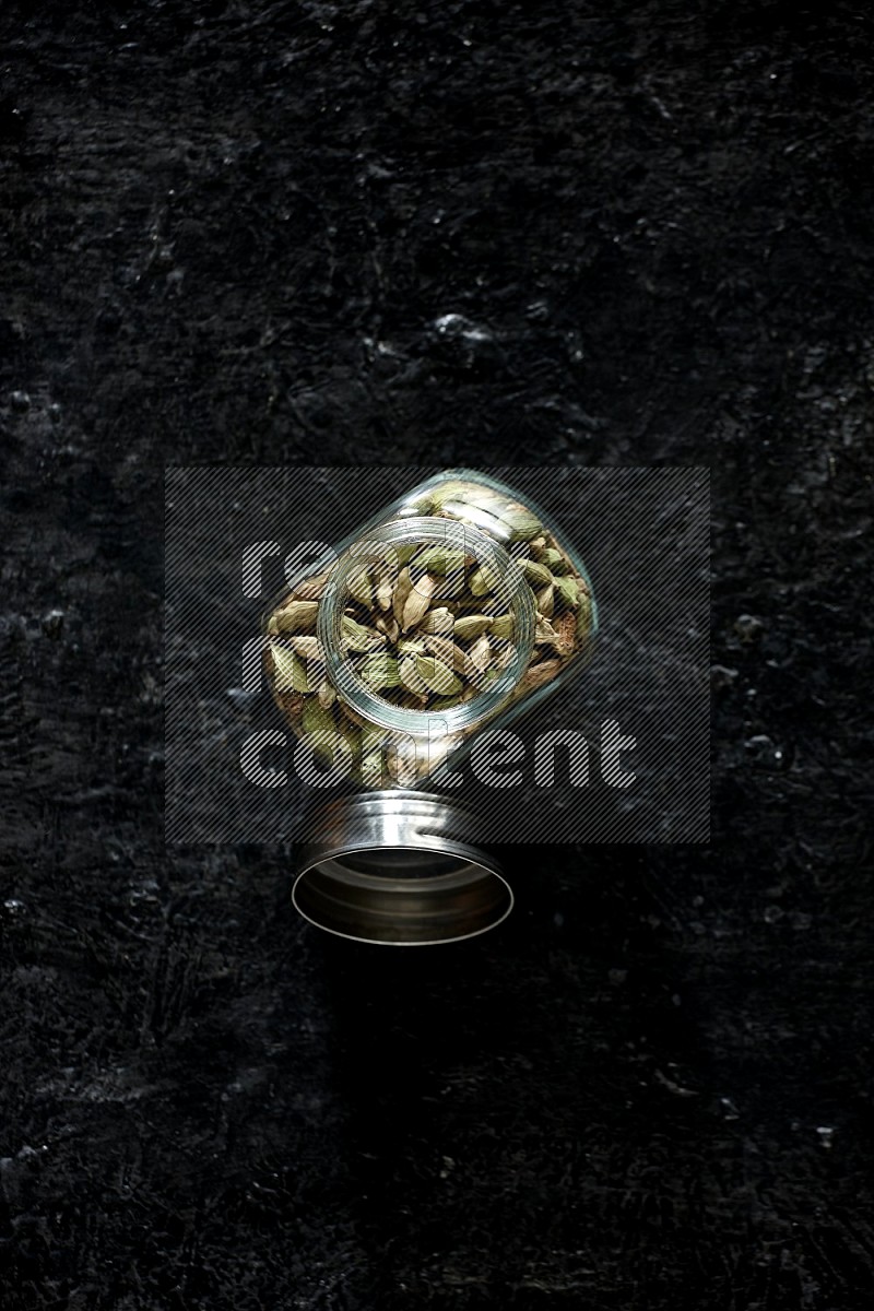 A glass spice jar full of cardamom seeds on textured black flooring