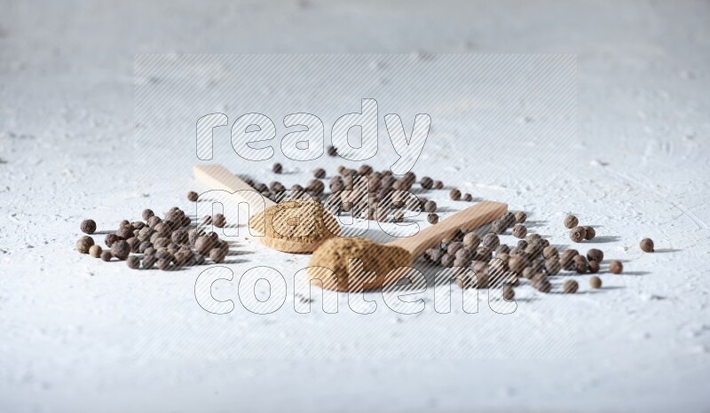 Wooden spoons full of allspice powder and allspice whole balls spreaded on a textured white flooring