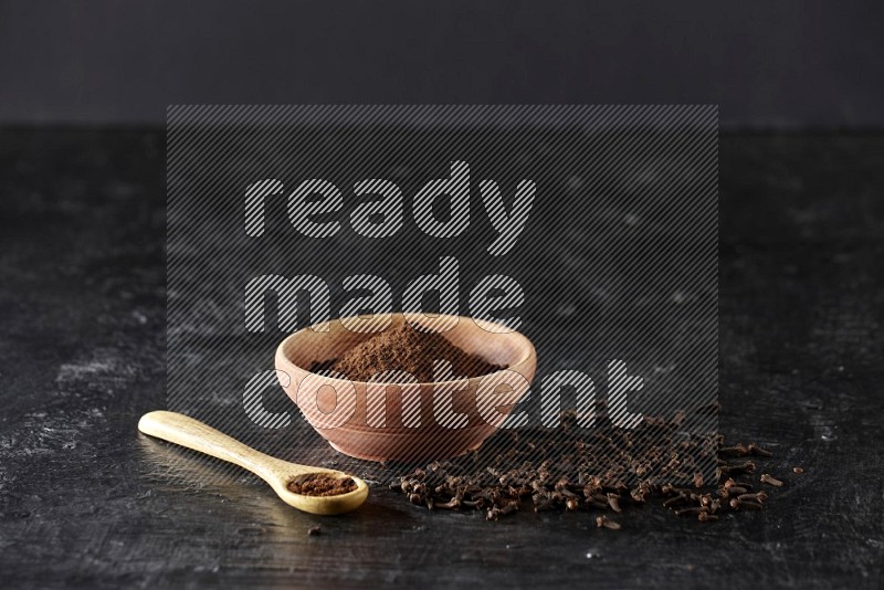 A wooden bowl and wooden spoon full of cloves powder with spreaded cloves on textured black flooring