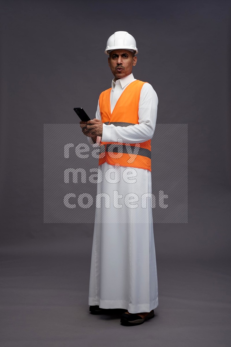 Saudi man wearing thob with engineer vest standing texting on phone on gray background