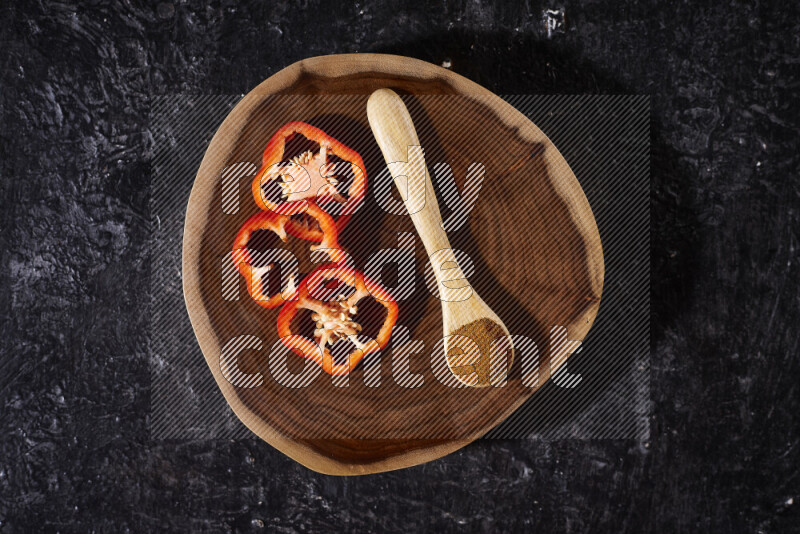 A wooden spoon full of ground paprika powder with red bell pepper slices beside it, all on a wooden tray on black background