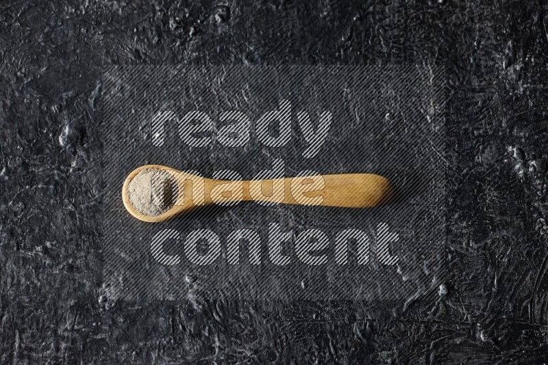 A wooden spoon full of white pepper powder on textured black flooring