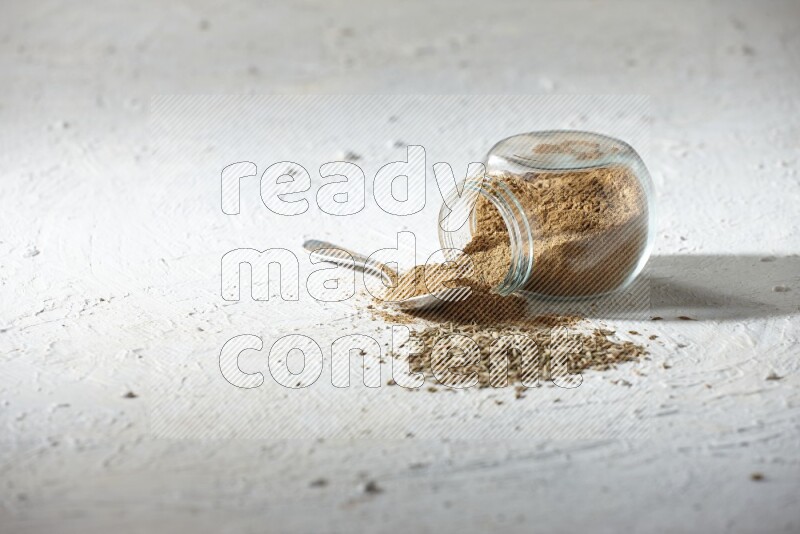 A glass spice jar and metal spoon full of cumin powder and the jar flipped and powder spilled out with cumin seeds on textured white flooring