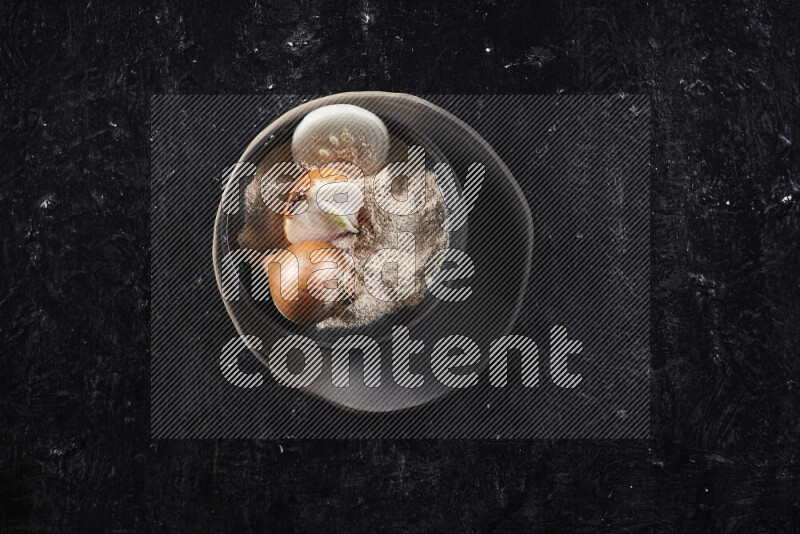 A black pottery bowl full of onion powder and some onion all on a pottery plate on black background