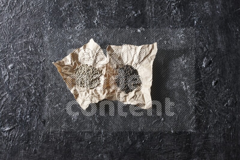2 Crumpled pieces of paper full of black and white pepper beads on a textured black flooring