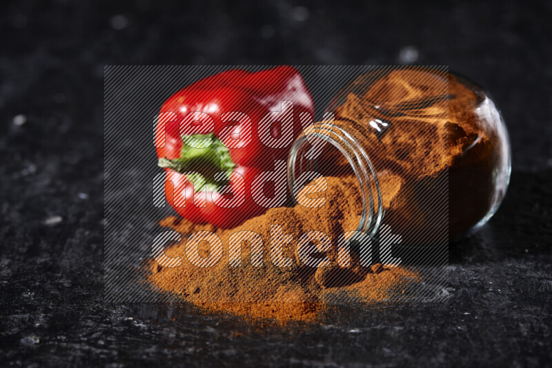 A glass jar full of ground paprika powder flipped with some spilling powder on black background