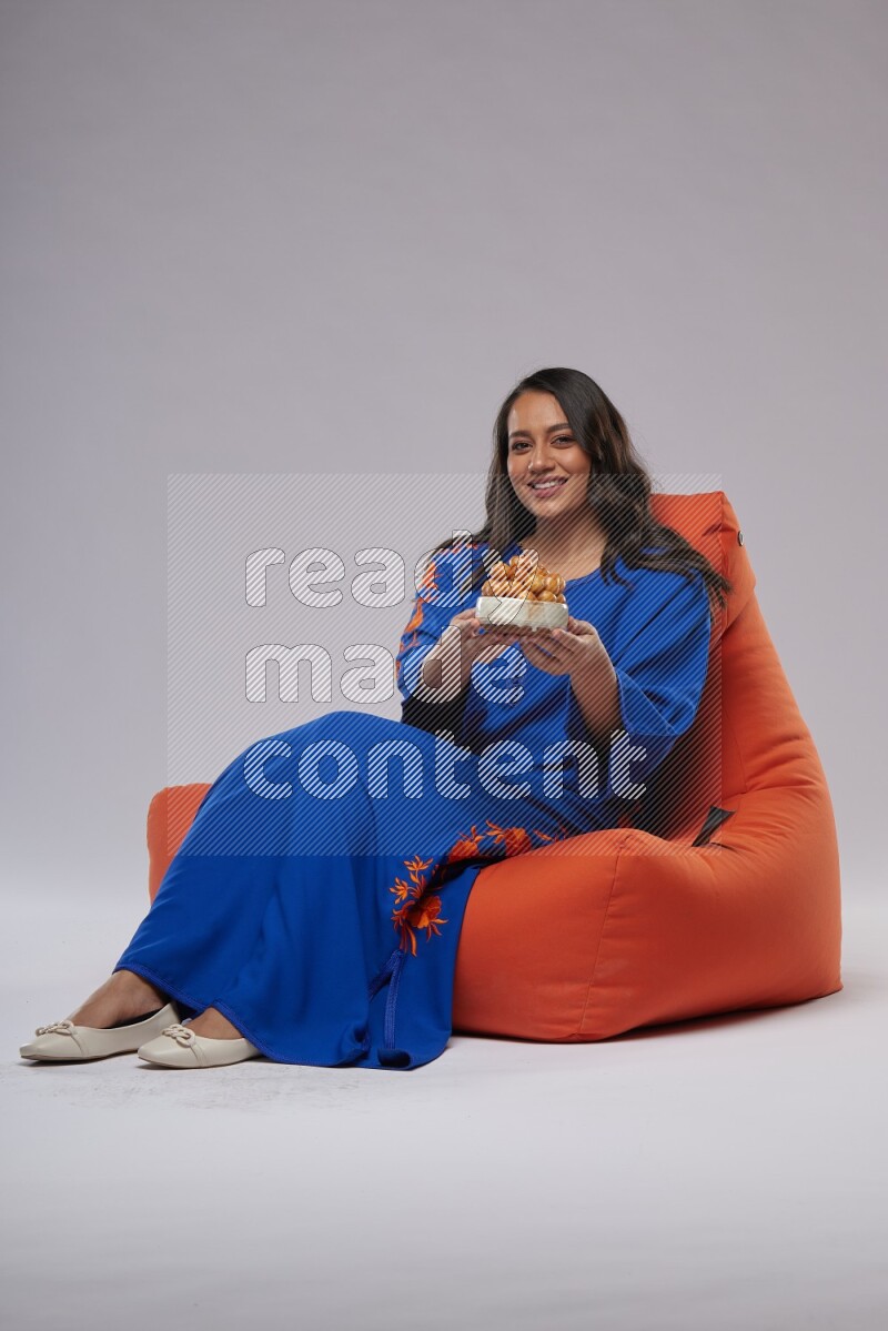 A Woman sitting on an orange beanbag wearing Jalabeya holding a plate of luqaimat