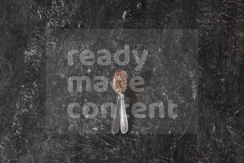 2 metal spoons full of flaxseeds on a textured black flooring