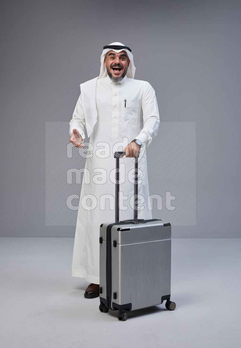 Saudi man wearing Thob and white Shomag standing holding Travel bag on Gray background