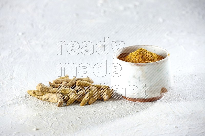 A beige pottery bowl full of turmeric powder and dried turmeric fingers beside it on textured white flooring