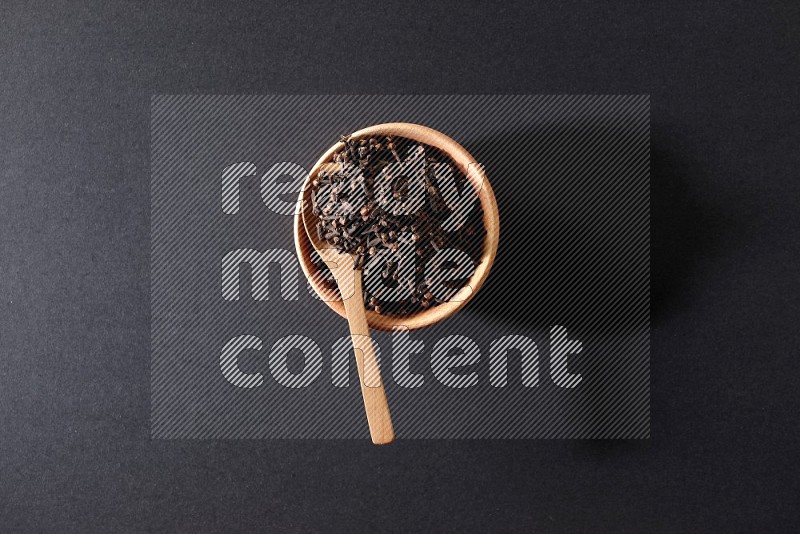 A wooden bowl and a wooden spoon full of cloves on a black flooring