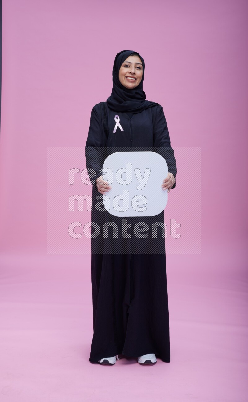 Saudi woman wearing pink ribbon on Abaya standing holding social media signs on pink background