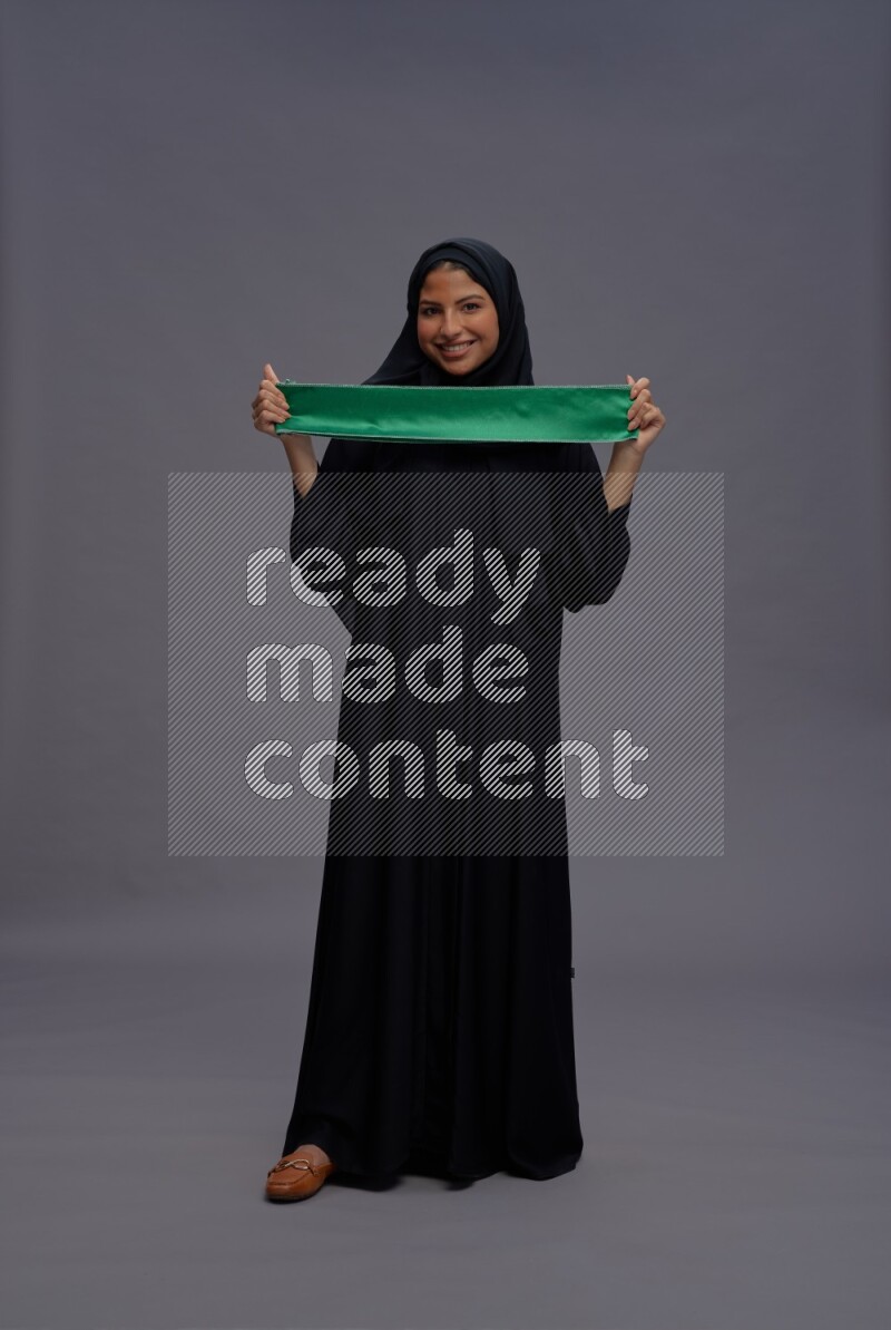 Saudi woman wearing Abaya standing holding Saudi flag on gray background
