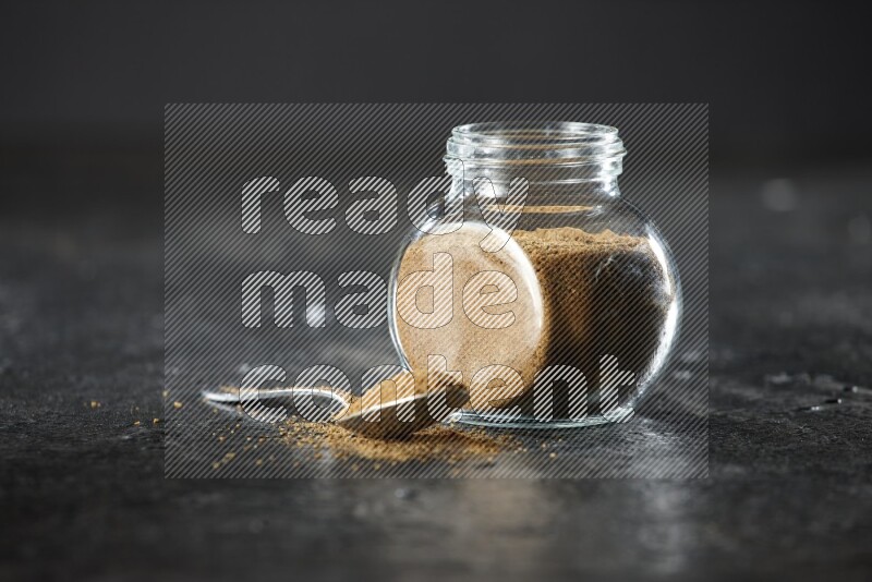 A glass spice jar and a metal spoon full of cumin powder on a textured black flooring