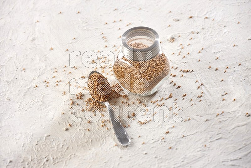 A glass spice jar and a metal spoon full of mustard seeds on a textured white flooring in different angles