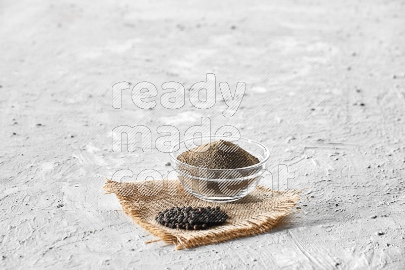 A glass bowl full of black pepper powder and black pepper beads on burlap fabric on textured white flooring