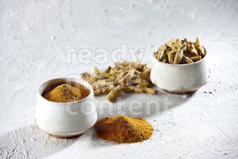 2 beige pottery bowls full of turmeric powder and dried turmeric whole fingers with powder and fingers next of it textured white flooring