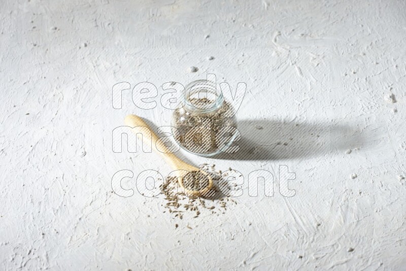 A glass spice jar and wooden spoon full of cumin seeds on textured white flooring