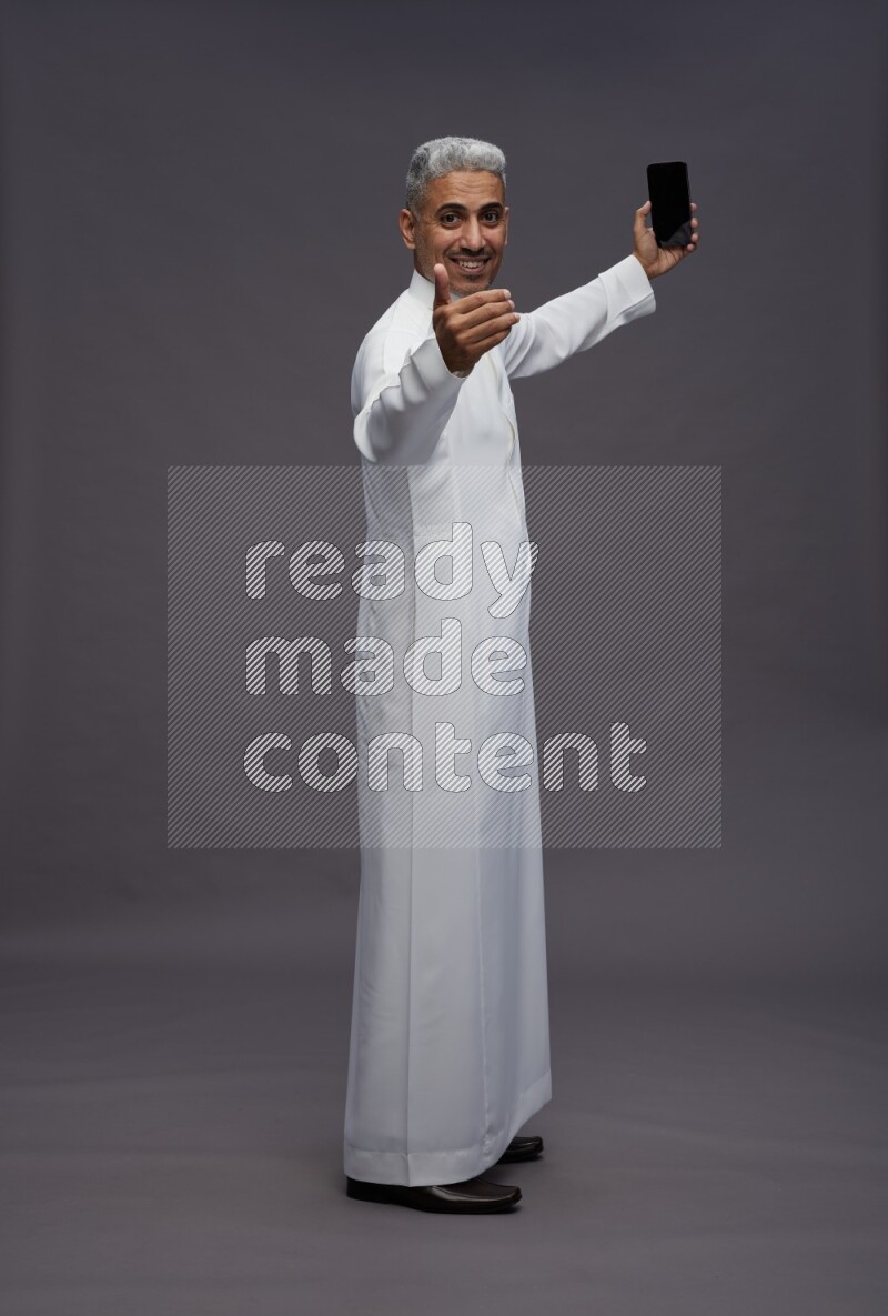 Saudi man wearing thob standing taking selfie on gray background