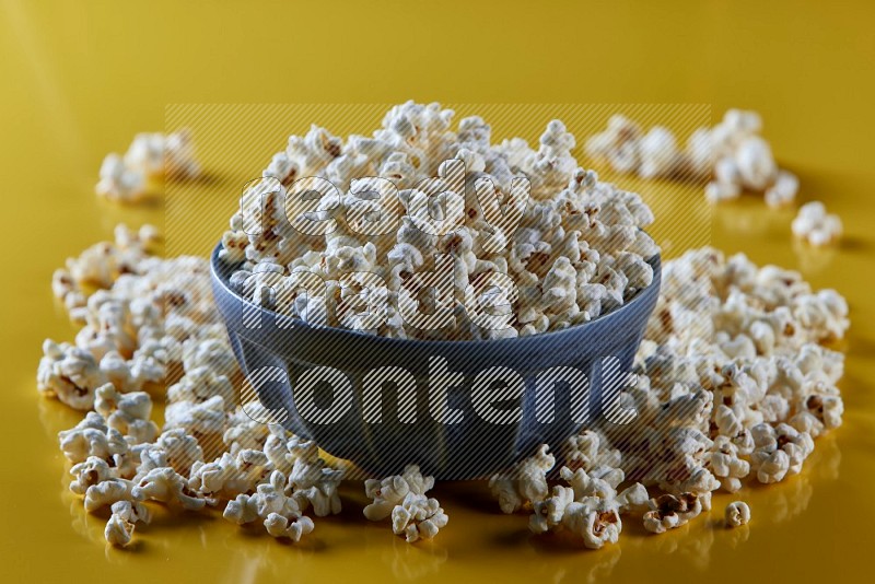 A blue pottery bowl full of popcorn with popcorn beside it on a yellow background in different angles