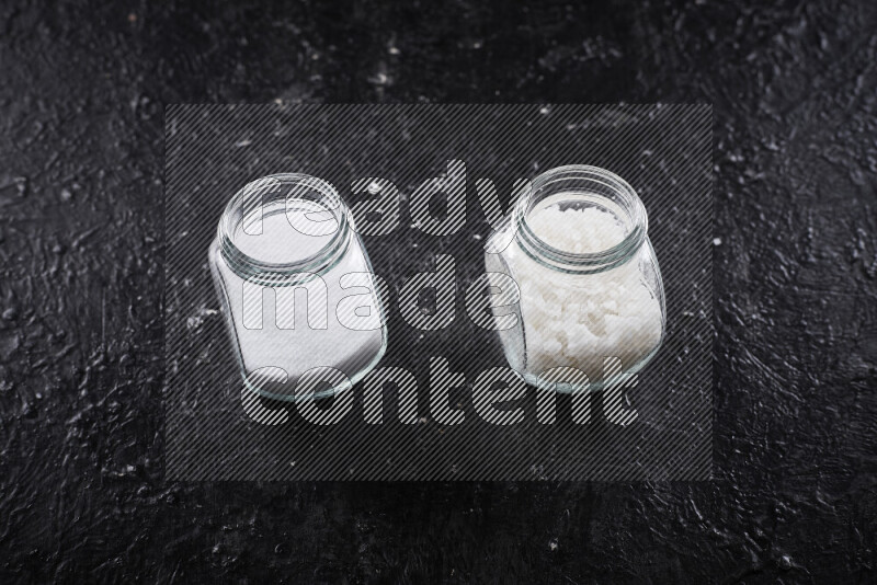 2 glass jars one is filled with table salt and the other with coarse sea salt on black background