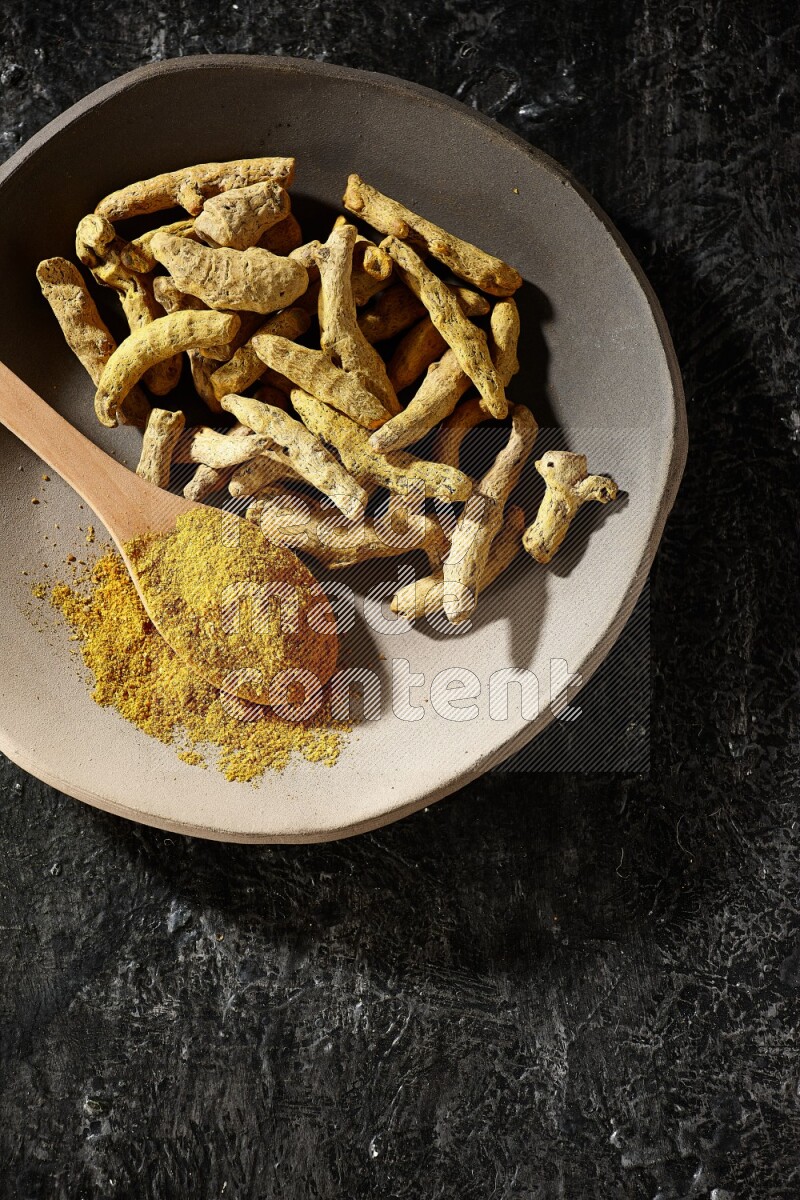 A plate filled with dried turmeric fingers and a wooden spoon full of turmeric powder on a textured black flooring