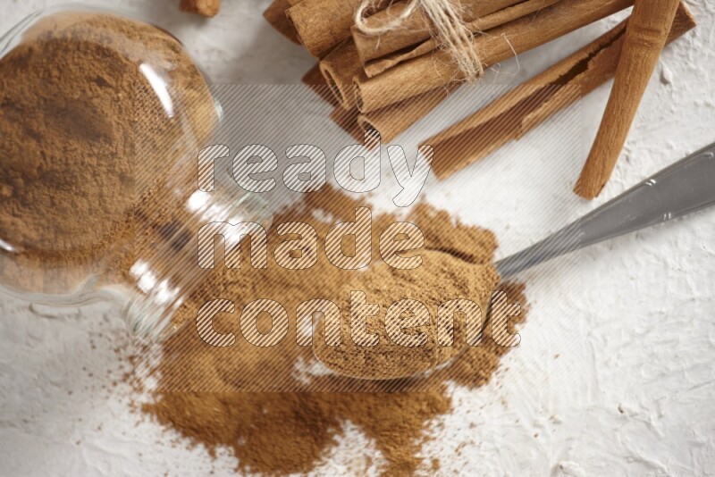 Flipped herbs glass jar full of cinnamon powder with a metal spoon full of powder and cinnamon sticks on a textured white background