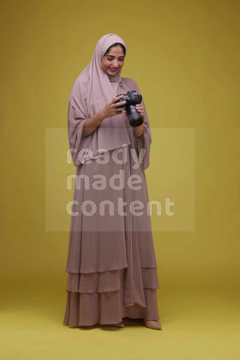 A woman Holding a Camera on a Yellow Background wearing Brown Abaya with Hijab