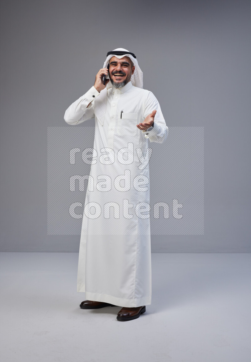 Saudi man Wearing Thob and white Shomag standing talking on phone on Gray background