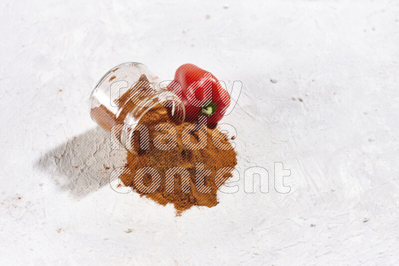 A glass jar full of ground paprika powder flipped with some spilling powder on white background