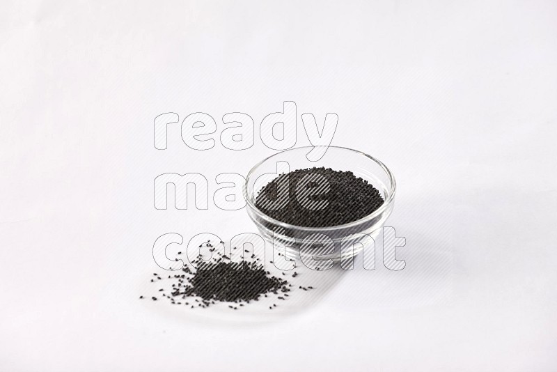 A glass bowl full of black seeds and some more seeds spread next to it on a textured white flooring
