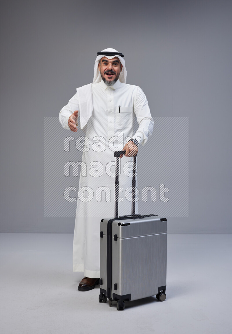 Saudi man wearing Thob and white Shomag standing holding Travel bag on Gray background