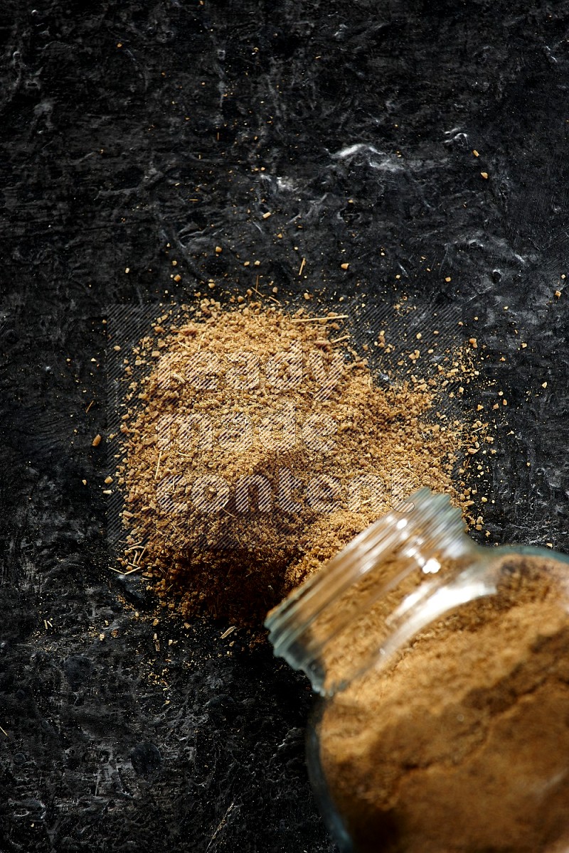 A flipped glass spice jar full of cumin powder and powder spilled out on a textured black flooring