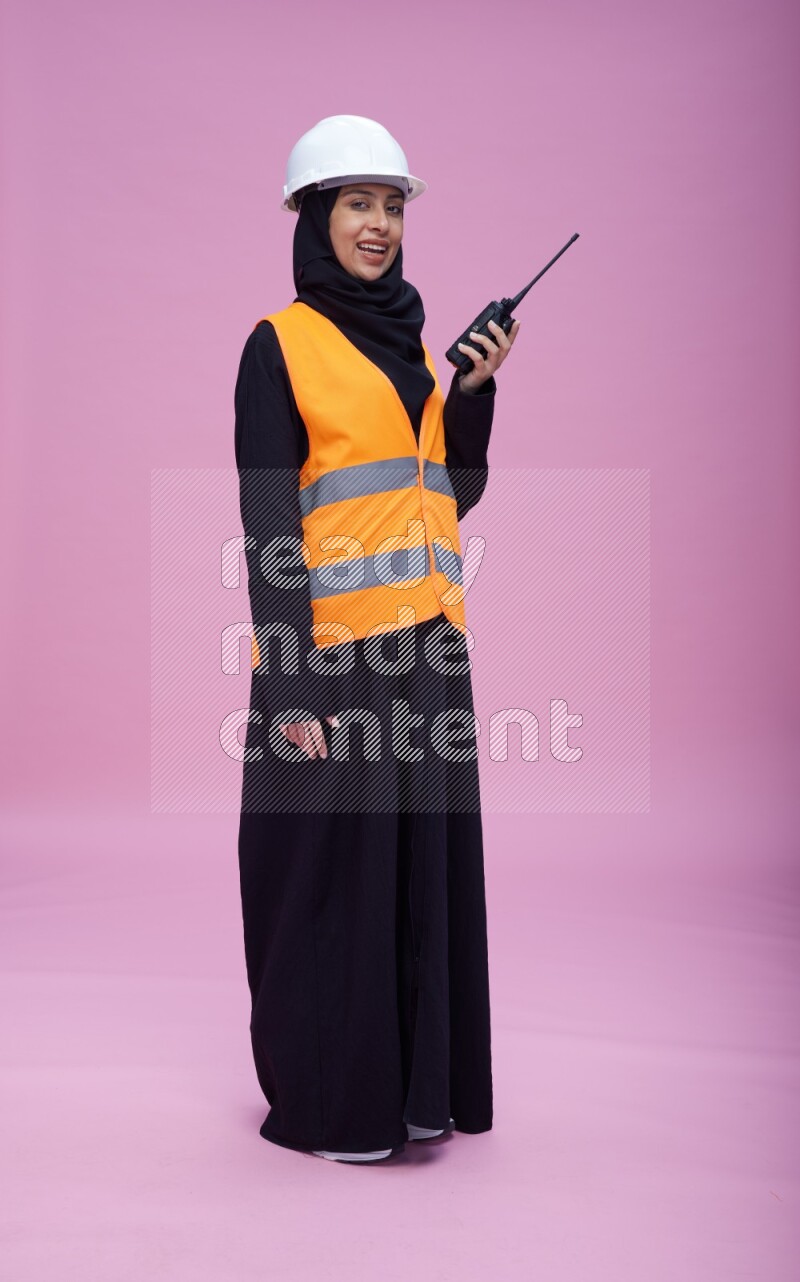 Saudi woman wearing Abaya with engineer vest and helmet standing holding walkie-talkie on pink background
