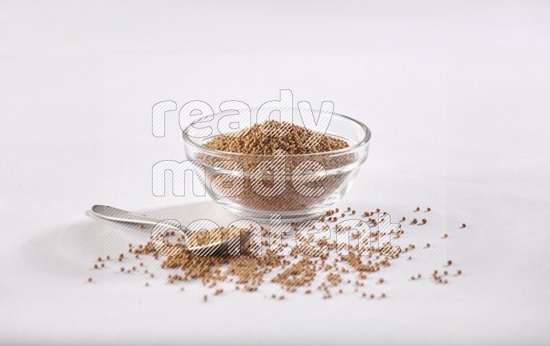 A glass bowl and metal spoon full of mustard seeds on a white flooring