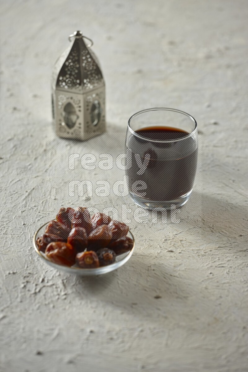 A silver lantern with different drinks, dates, nuts, prayer beads and quran on textured white background
