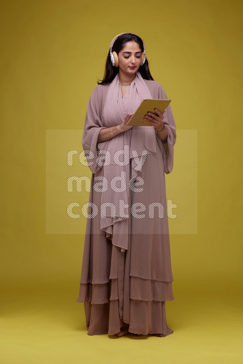 A woman Holding an Ipad with Headphone on a Yellow Background wearing Brown Abaya