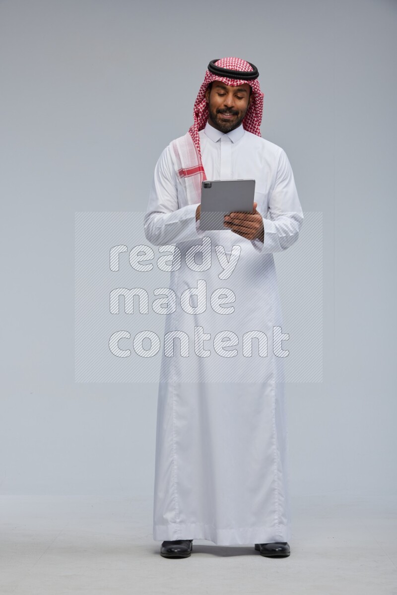 Saudi man Wearing Thob and shomag standing working on tablet on Gray background