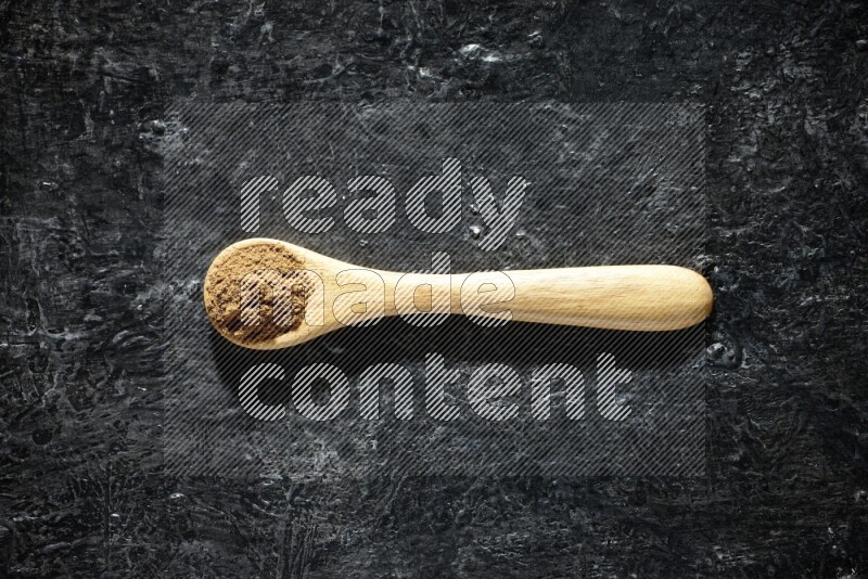 A wooden spoon full of allspice powder on a textured black flooring