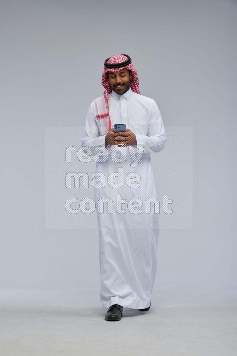 Saudi man Wearing Thob and shomag standing texting on phone on Gray background