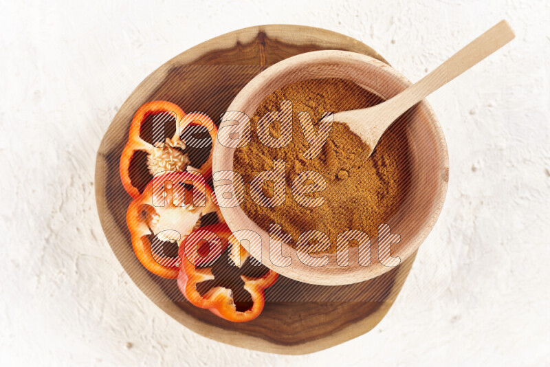 A wooden bowl full of ground paprika powder and sliced red bell pepper beside it all on a wooden tray on white background