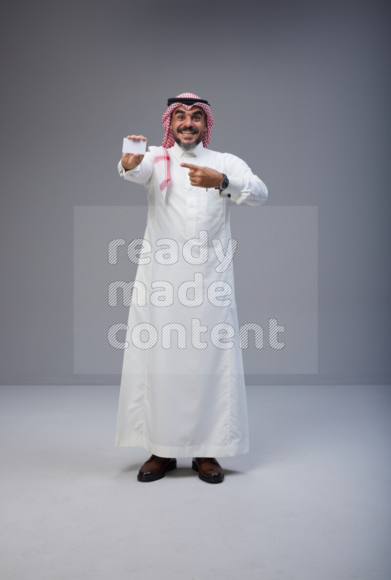 Saudi man Wearing Thob and red Shomag standing holding ATM card on Gray background