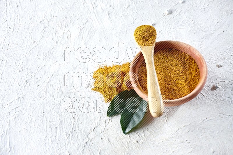 A wooden bowl and wooden spoon full of turmeric powder on textured white flooring