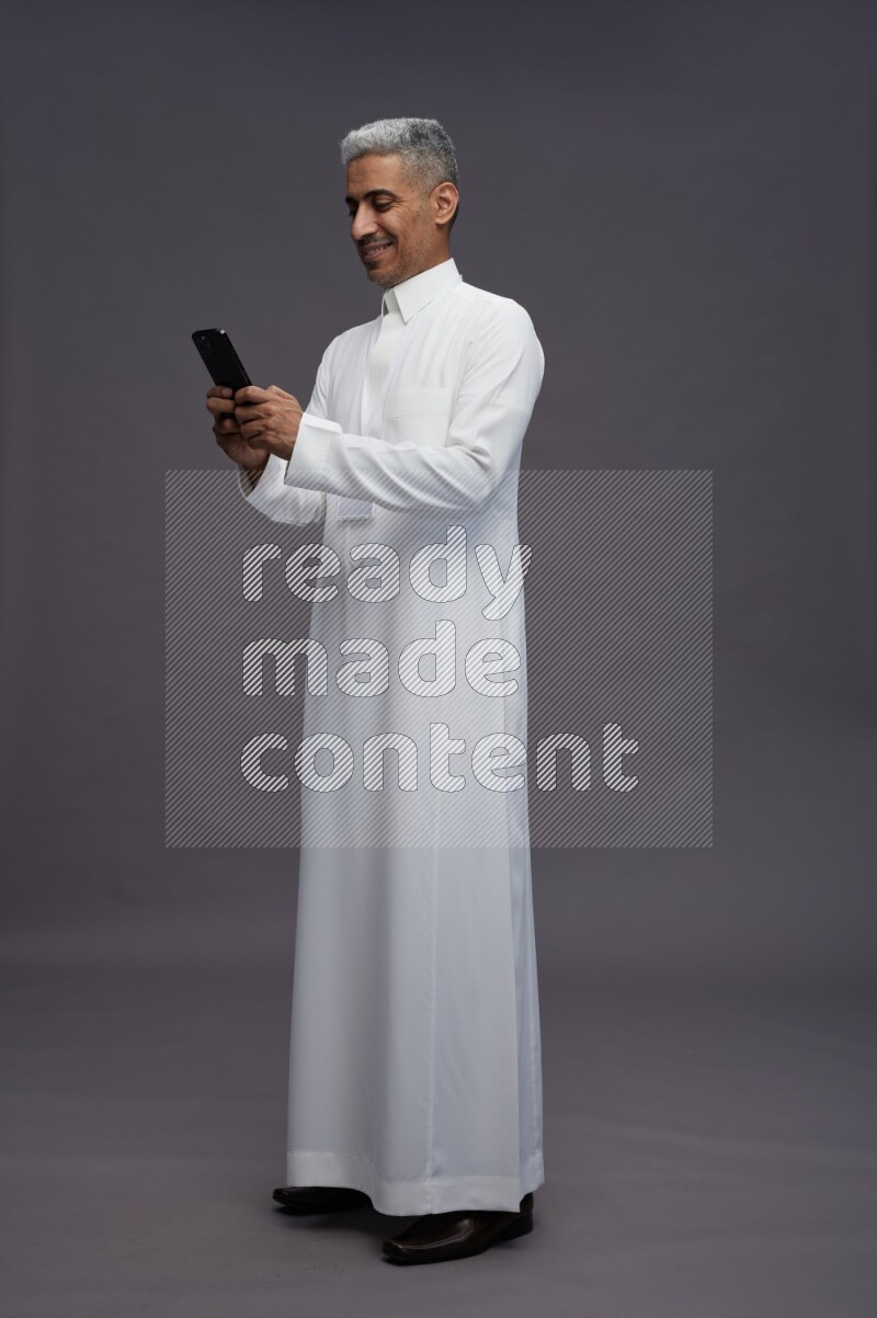 Saudi man wearing thob with neck strap employee badge standing texting on phone on gray background