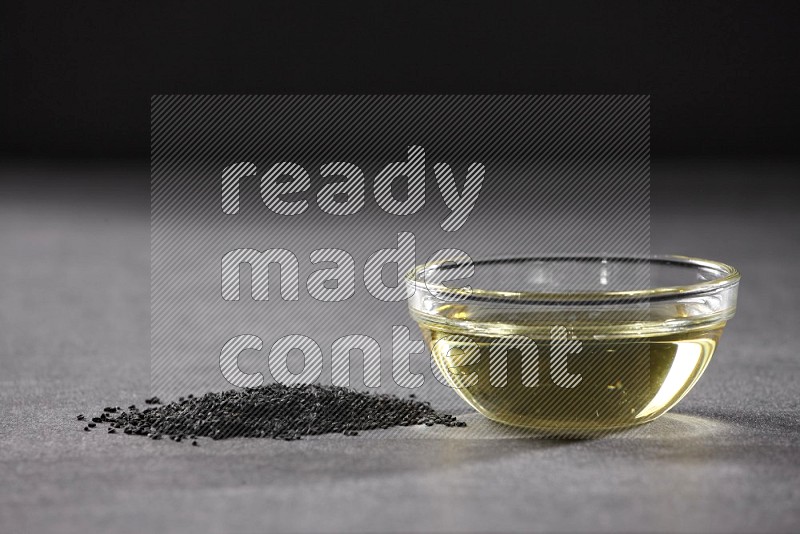 A glass bowl full of black seeds oil and black seeds beside it on a black flooring