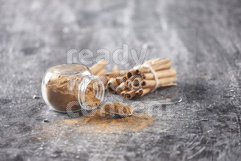 Herbal glass jar full cinnamon powder flipped and a metal spoon full of powder, cinnamon sticks stacked and bounded in the back on textured black background in different angles